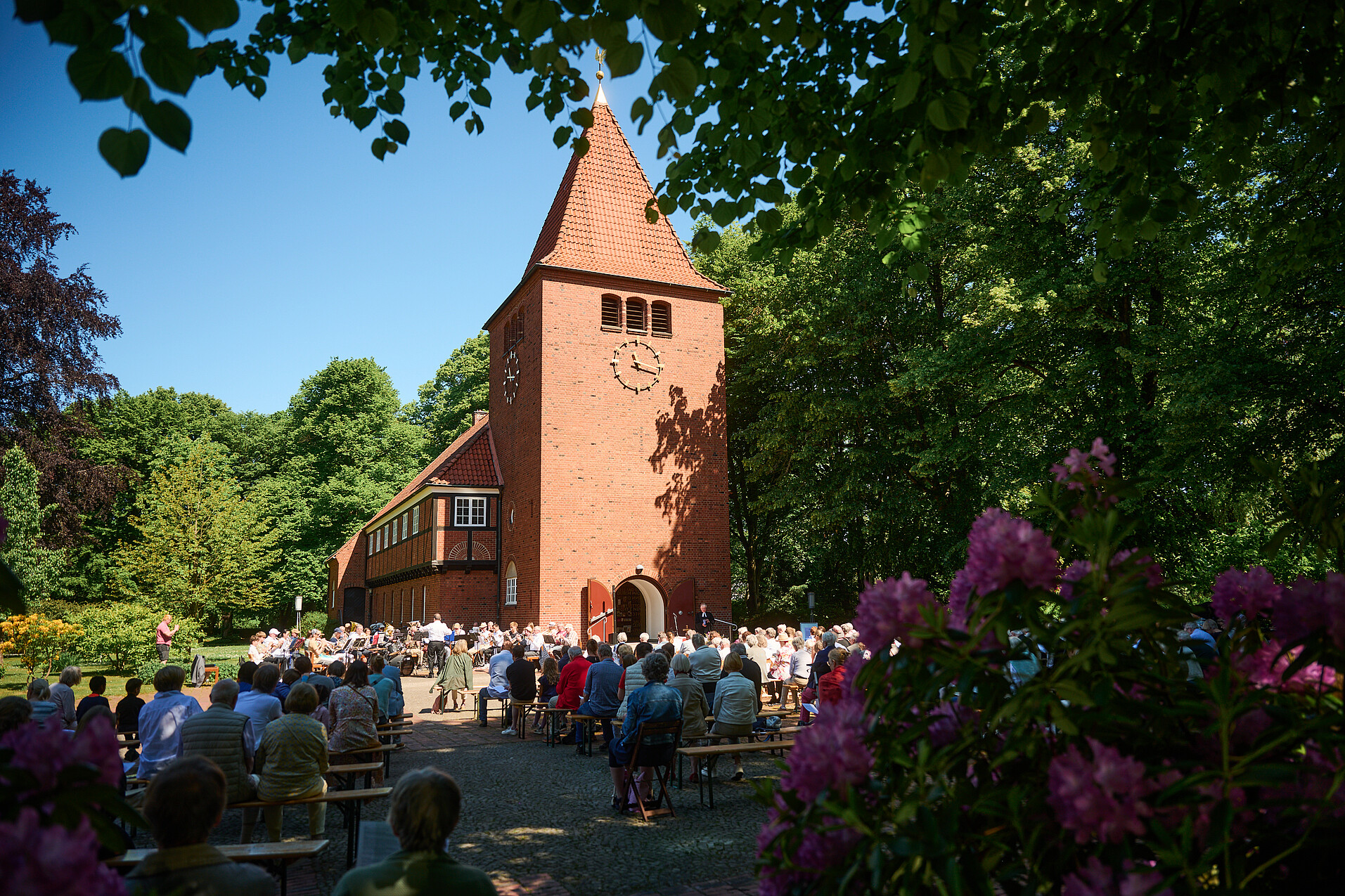 Kirche vor blauem Himmel. Eine Gemeinde sitzt auf Bänken. Es spielt ein Posaunenchor.
