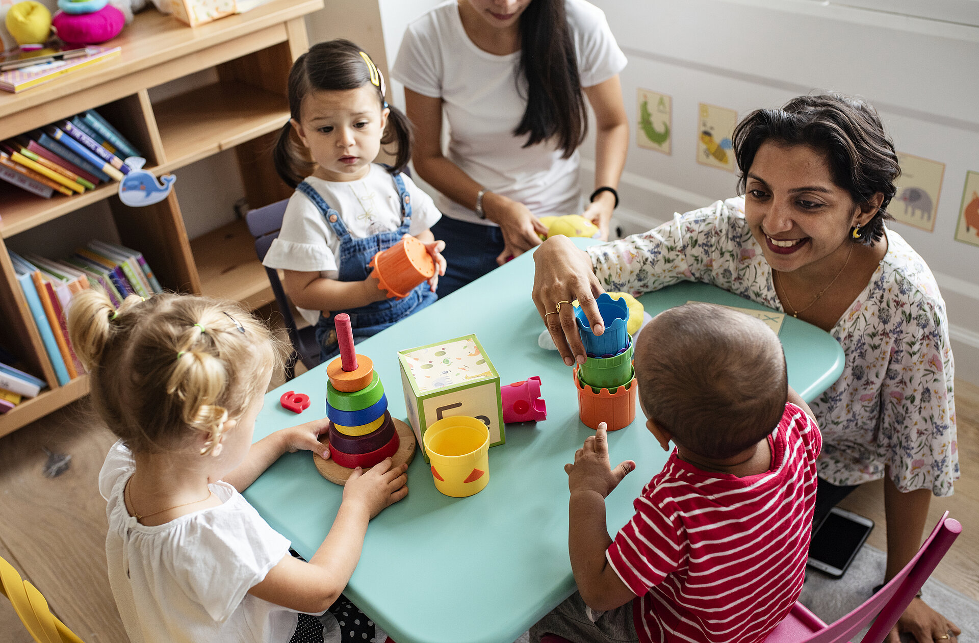 Drei Kinder und zwei erwachsene Frauen sitzen gemeinsam an einem Tisch in der Spielecke und spielen mit Spielzeugbechern zum Stapeln
