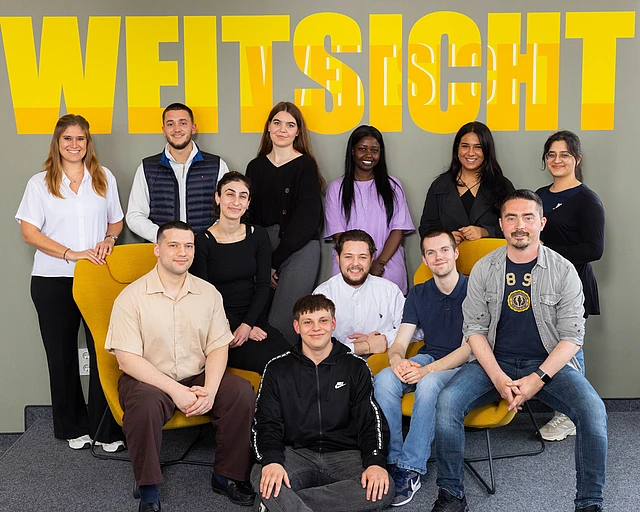 Gruppenfoto mit Ausbildenden und einem Ausbildungsleiter vor einer Wand mit dem gelben Schriftzug Weitsicht