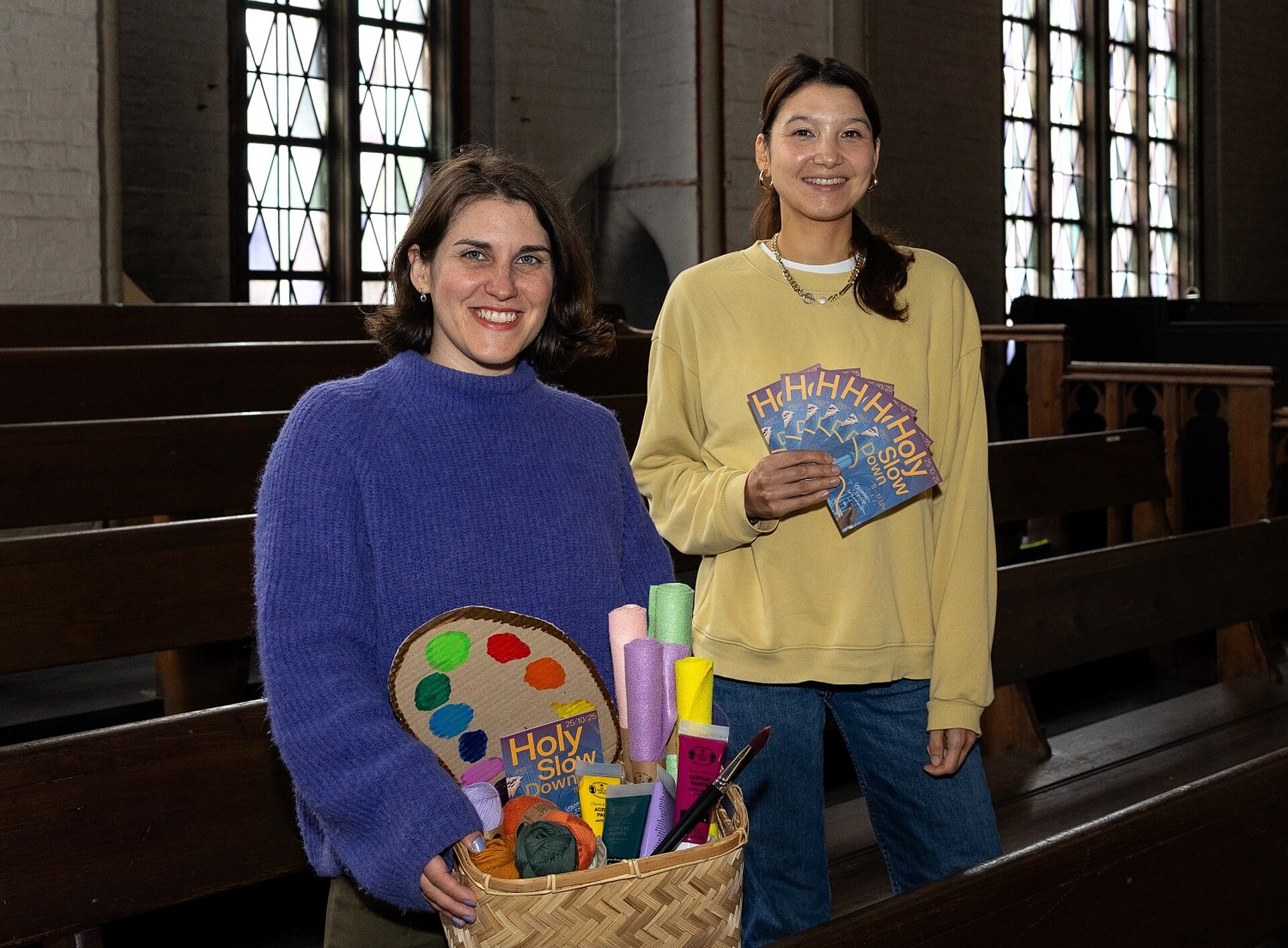 Zwei Frauen stehen in einer Kirche. Eine hält einen Korb mit Bastelmaterialien und eine Flyer, auf denen Holy Slow Down steht.